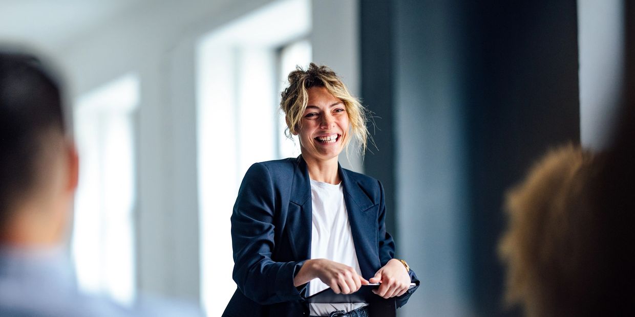 A smiling woman in a blazer engaging with others in a bright room.