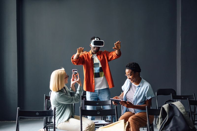 An African American male is standing up and wearing virtual reality goggles. He is making hand gestures in air. Two of his female friends are taking photos of him and smiling.