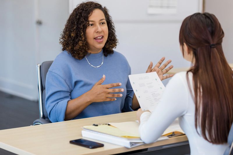 The mid adult woman gestures as she interviews for a job with an unrecognizable female manager.