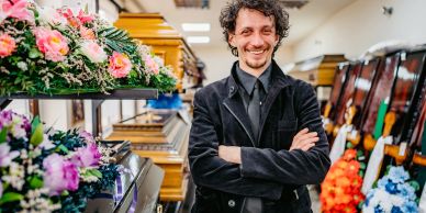 Man smiling in a funeral home among coffins and floral arrangements.