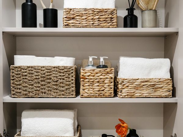 Organized shelves with wicker baskets, white towels, toiletries, and a small flower vase in a bathroom cabinet.