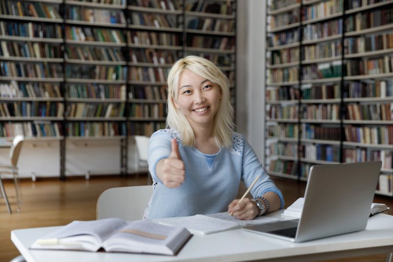 Attractive 20s Asian student girl shows thumbs up seated at table in library, holding pencil making exercise using laptop feel happy by effective and interesting study in university. Education concept