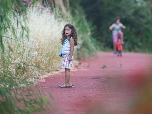 A young girl stands on a red path surrounded by greenery and flowers.