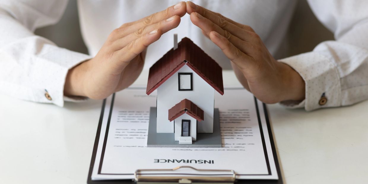 Hands protect a small house model on an insurance document.