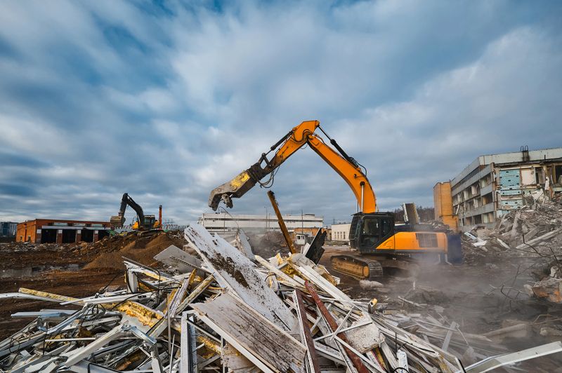 Yellow excavator destroyer with hydraulic scissors cuts reinforced concrete waste at abandoned industrial complex demolition site