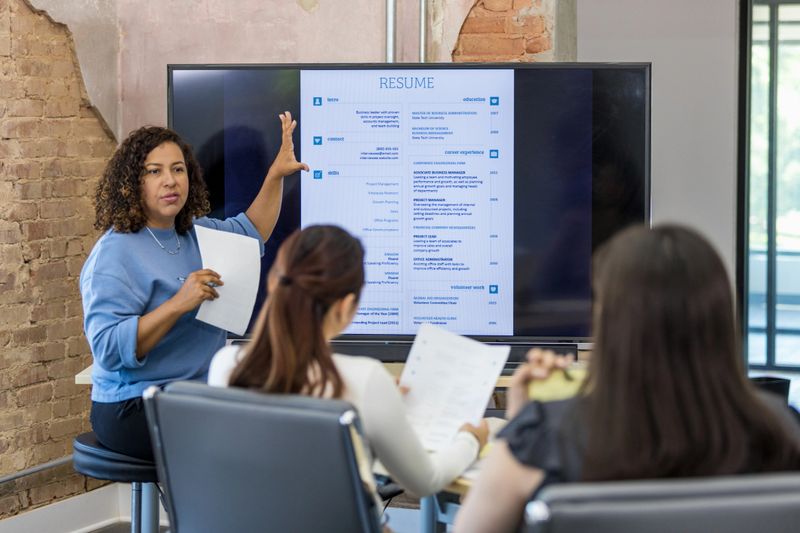 A female mid adult human resources representative leads a resume building class.  She uses a large projection screen.
