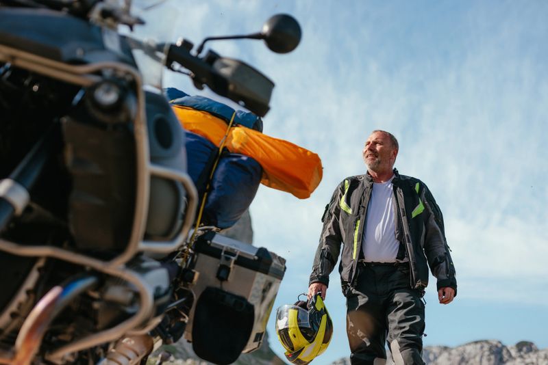 A smiling motorcycle rider and his bike are in a scenic mountainous landscape enjoying the nature.