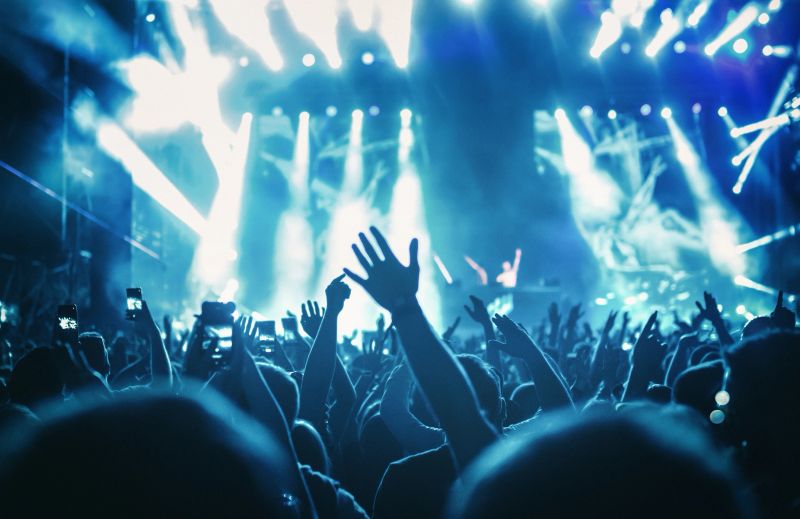 Rear view of large group of people enjoying a concert performance. There are many hands applauding and taping the show. Multi colored lasers and spot lights firing from the stage.Silhouettes have been significantly liquified.
