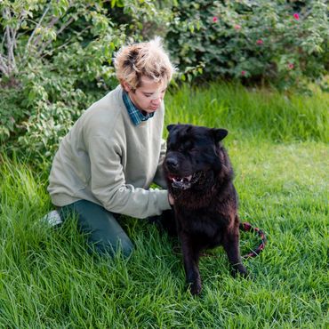 Person with blonde hair kneeling on grass, petting a large black dog.