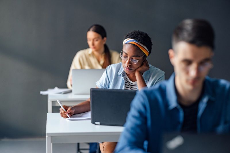 Serious African-American woman sitting at desk with laptop computer and writing notes in a notebook  during lecture at college.