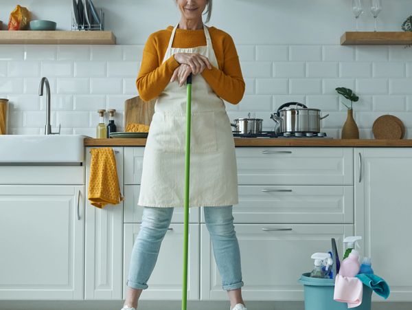 Smiling woman in apron holding mop in a clean modern kitchen.