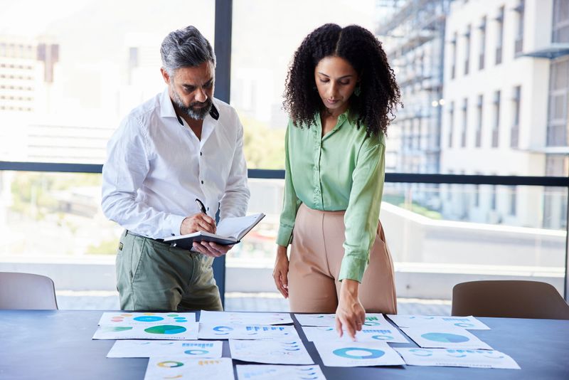 Two diverse businesspeople going over charts and graphs on paper at a table in the boardroom of an office