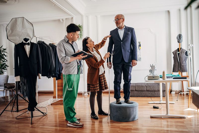 Female tailor taking measurements on a mature male client for a new suit, while talking to a male fashion designer.