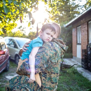 A soldier holds a sad child while standing outside near cars and a house.
