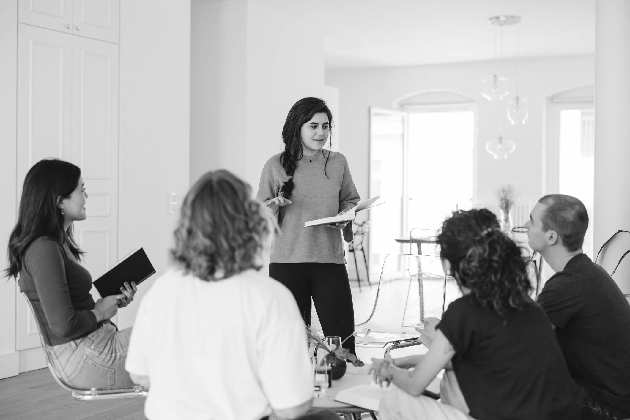 A young woman stands speaking to a seated group in a bright room.