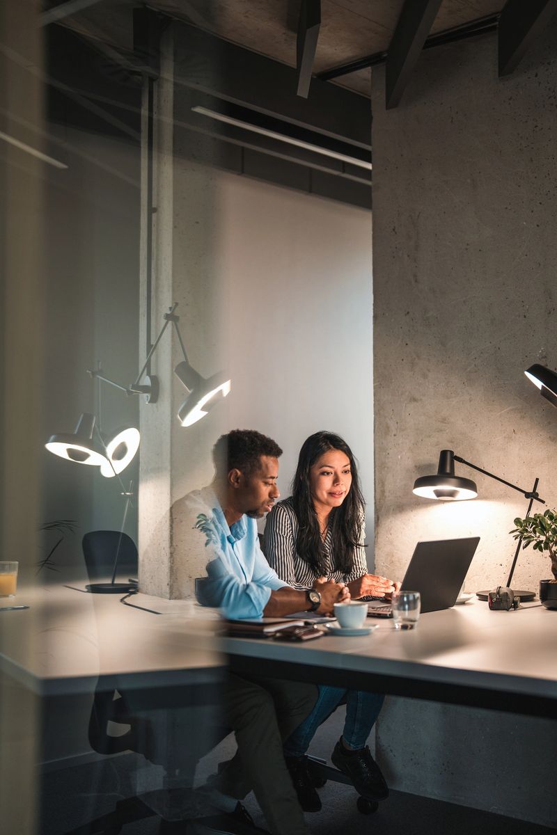 Candid shot of African American male and Hispanic female working hard at the office during the night. They are seated in a desk and their glass door is open reflecting the light from their desk lamp.