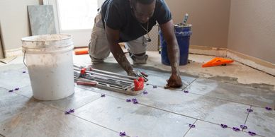 A man is installing floor tiles with tools and buckets around him.