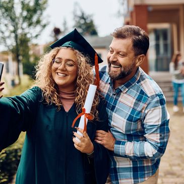 Young graduate in cap and gown takes a selfie with a happy man outdoors.