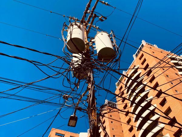 Electrical pole with tangled wires against clear blue sky near tall buildings.