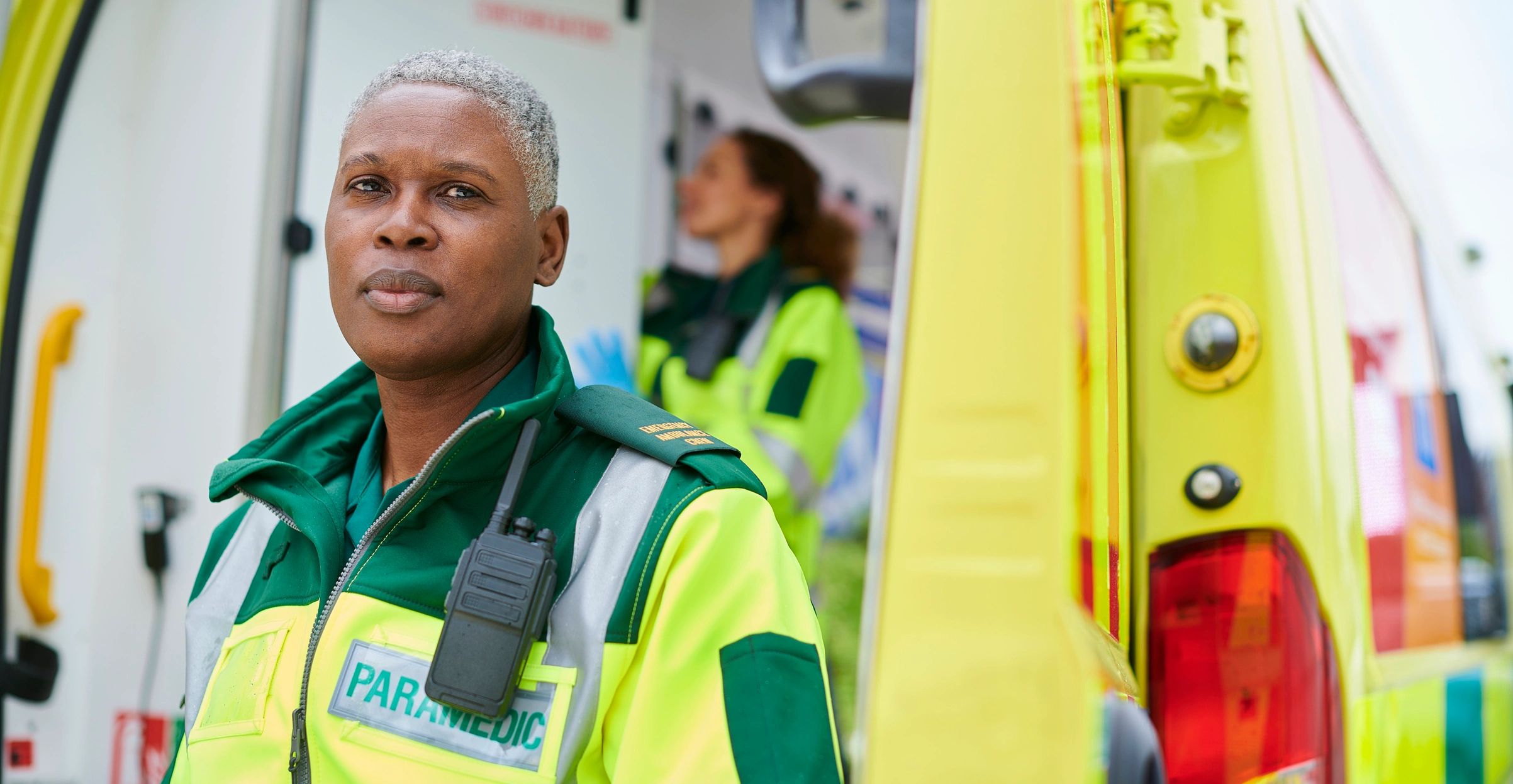 A paramedic in their uniform looking straight to camera and an ambulance behind them