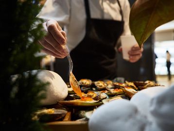 Chef drizzling sauce over cooked mussels in a kitchen setting.