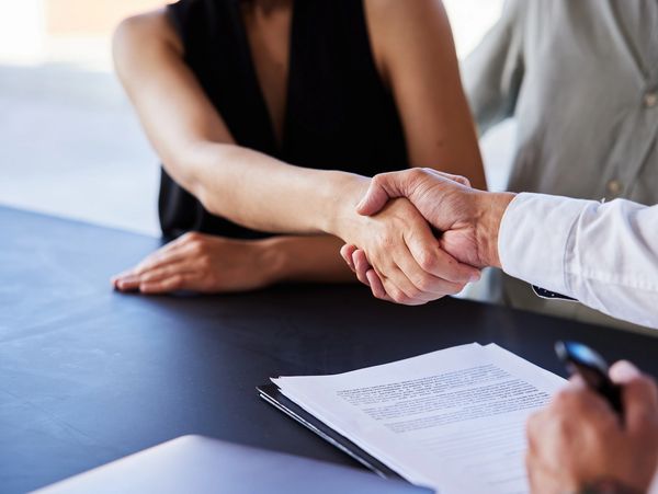 Two people shaking hands over a contract on a desk.