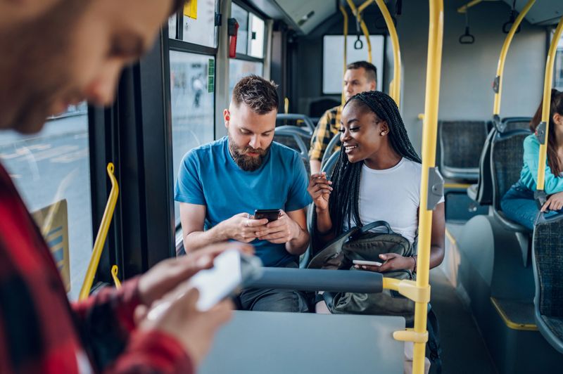Happy multiracial couple of friends sitting in a bus and using a smartphone to message their friends who they are going to meet. Public transportation. Diverse romantic couple riding a bus.