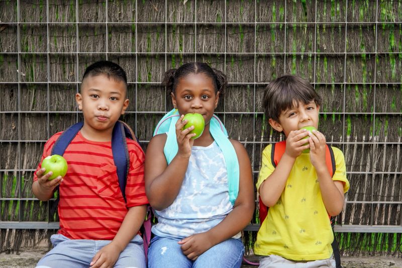 multiethnic children with backpacks eating apples at school entrance