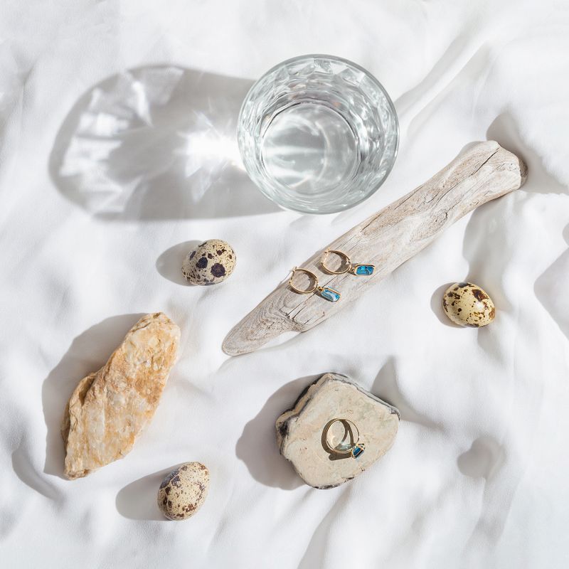 Abstract creative composition made of elegant jewellery set of golden ring and earrings with blue topaz on white background with stones, wood,quail eggs and a glass of water.  Minimal style composition. selective focus