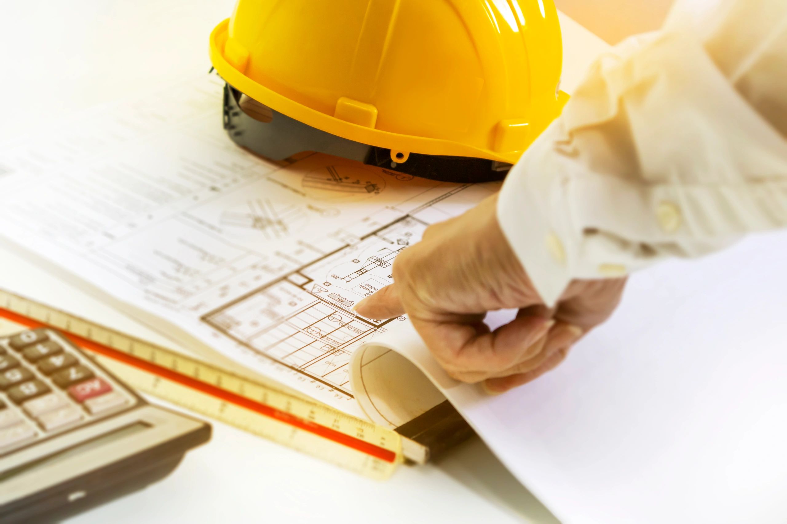 An engineer points at architectural plans beside a yellow hard hat and calculator.