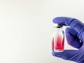 Gloved hand holding vial with red liquid against white background.