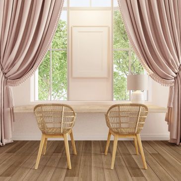 Cozy workspace with two wicker chairs, a wooden desk, and soft pink curtains framing large windows.