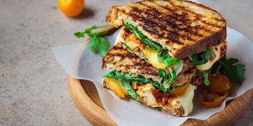 Grilled sandwich stacked with fresh greens, melted cheese, and tomatoes on a wooden plate.