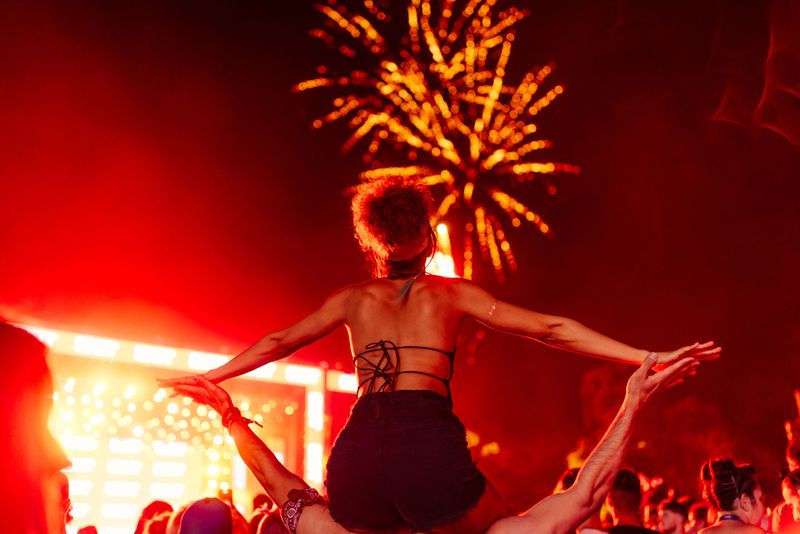 Man carrying his girlfriend on shoulders and watching fireworks at music festival. View from behind.