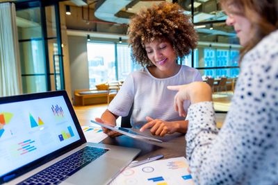 Two women discussing data charts on a tablet and laptop in a modern office.