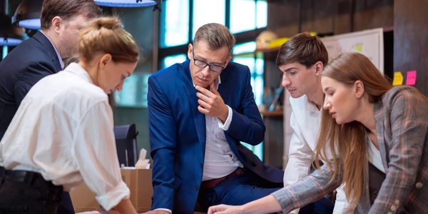 A team of five professionals collaborating over documents in a modern office, delivering expert business consulting services that enhance business development, provide organizational coaching, streamline operations for growth, and support clients in obtaining business funding