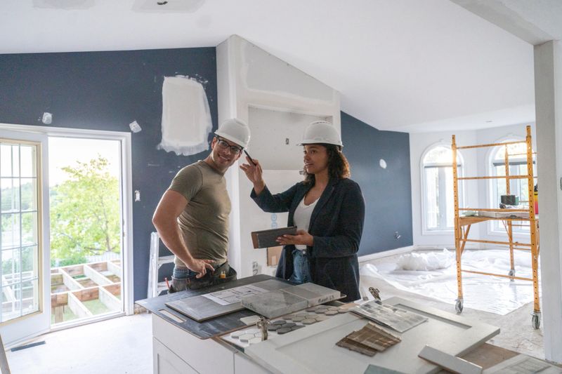 A young couple stand together in the kitchen as they try to make decision on design elements for their renovation.  They are both dressed casually and have hardhats on as they look through the plans together and confirm measurements and design details.