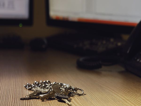 A bunch of keys on a wooden desk with computer monitors in the background.