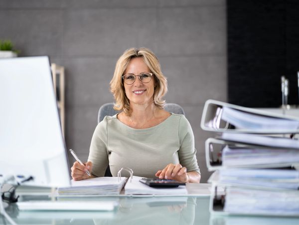 Smiling woman working at desk with stacks of binders and documents.