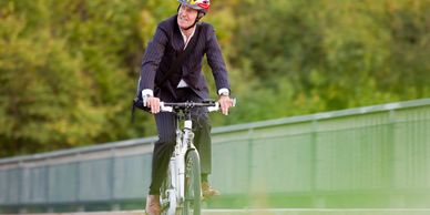 Man in business suit cycling with a helmet on a path beside greenery.