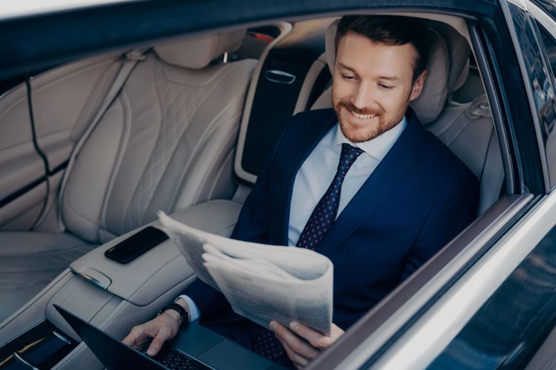 Relaxed male entrepreneur dressed in blue suit, reading latest newspaper and checking news regarding his successful company, while working on notebook, taking ride to work