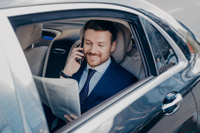 Elegant businessman in formal stylish wear traveling in luxury car,reading newspaper and searching financial information while talking on mobile phone, smiling with satisfaction to hear good news