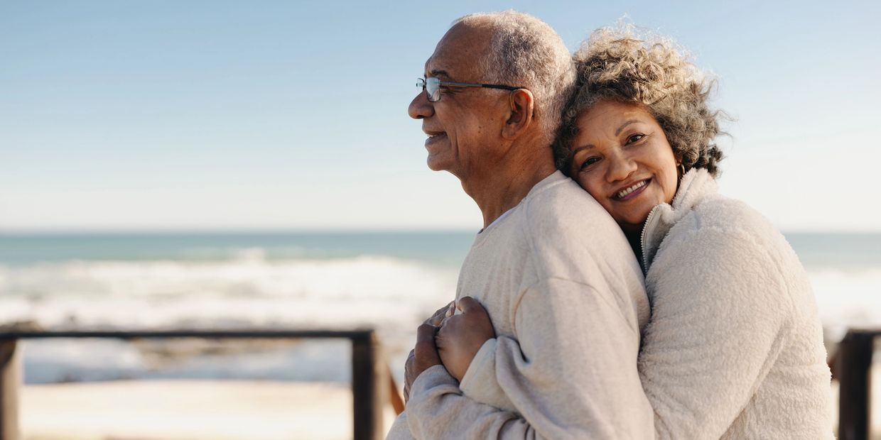 a woman holding her husband as they consider medicare options while enjoying the ocean scenery