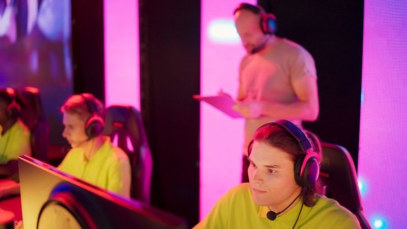 Diverse gamer group with african ethnicity players with a coach playing competitive computer game on a stage. Wearing headset and talking into the microphone to communicate with the yellow pro team.