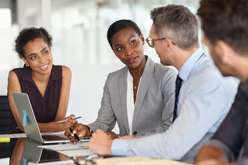 Successful business people working and discussing charts and graphs with laptop in conference room. Mature african businesswoman sharing progress on project to leader in modern office. Multiethnic business team checking new startegy on project.