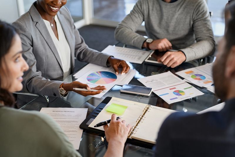 High angle view of businessmen and business women discussing stockmarket charts in office. Businesspeople discussing work at conference table in office with staff. African american leader working and discussing with colleagues in conference room during brief.