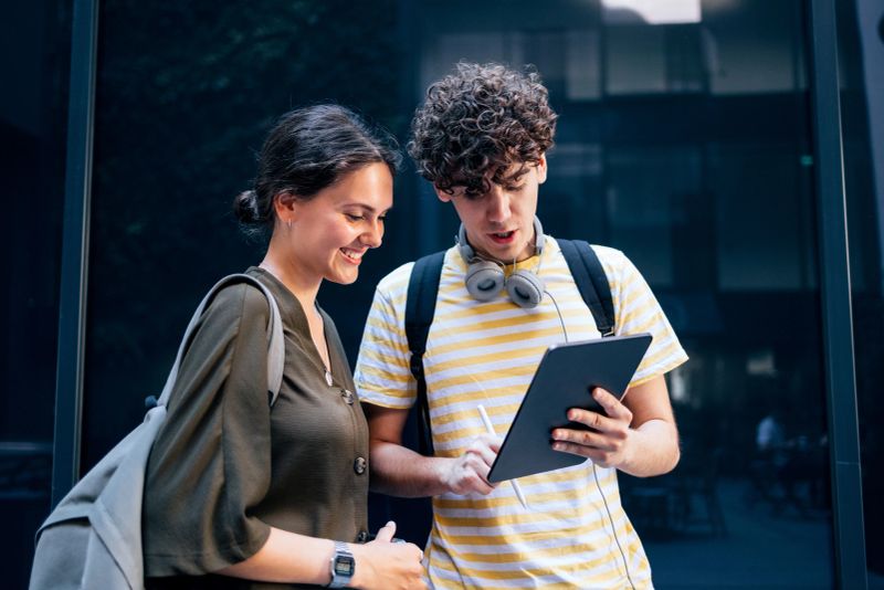 A couple of cheerful students outdoors, standing next to each other. Them man is using the digital tablet and drawing something on it.