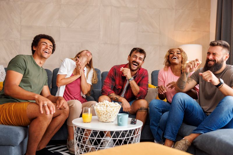 Group of friends eating popcorn and laughing while watching funny movie on TV at home