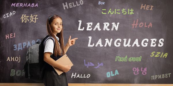 Young girl with a backpack pointing at 'Learn Languages' on a chalkboard with greetings in various languages.