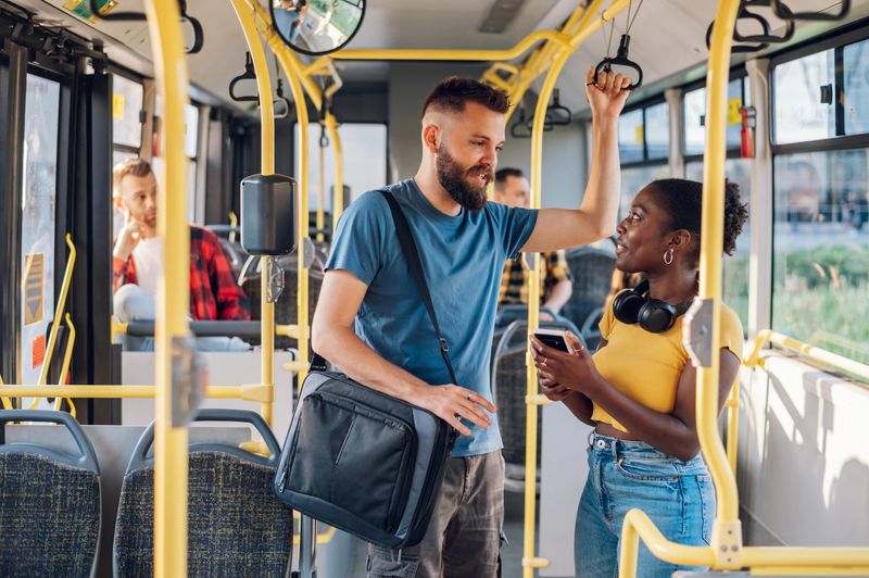 Cheerful diverse friends smiling and talking together and using a smartphone while riding a bus in the city and going to work. Standing in public transport, commuting by bus. Diverse romantic couple.